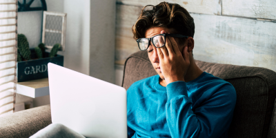 A person wearing glasses and a blue shirt sits on a couch with a laptop, resting their head on one hand.