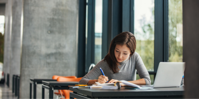 A woman is sitting at a table by a large window, writing in a notebook. A laptop, phone, and a pile of books are on the table.